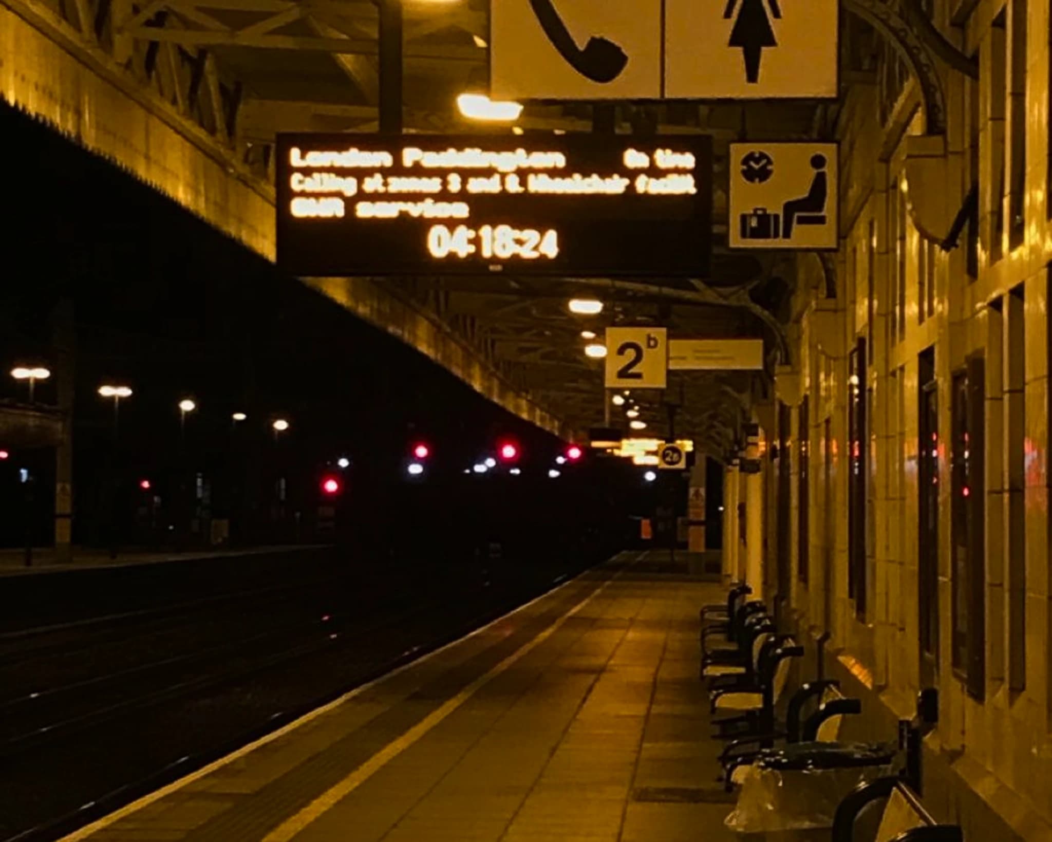 A near-empty train platform at night — a destination board reading “London Paddington”, benches along the wall