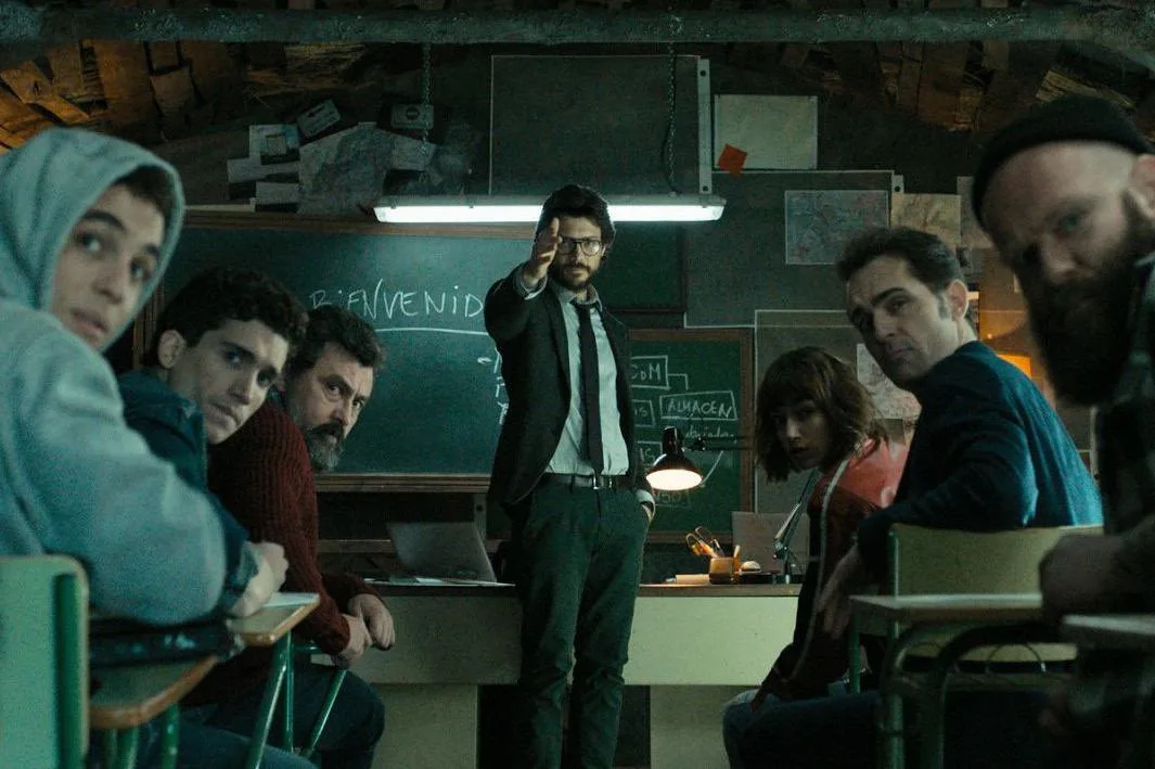 A still from the TV show Money Heist (La Casa de Papel) — the Professor standing before his crew gathered around desks in their planning room.